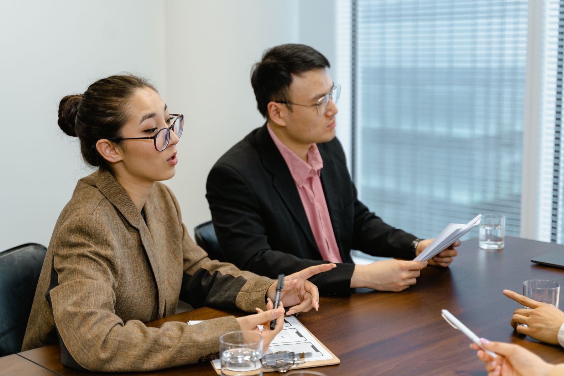 Two colleagues at a conference table, one explaining, the other holding papers.